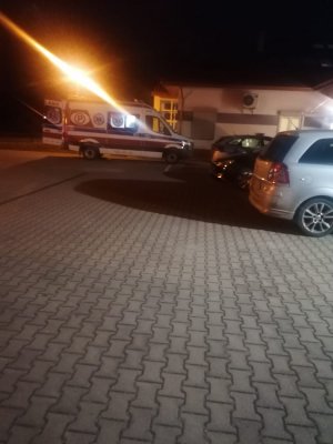 Pora nocna. w głębi karetka pogotowia z otwartymi tylnymi drzwiami stojąca na parkingu przed budynkiem. Z prawej zaparkowane samochody osobowe