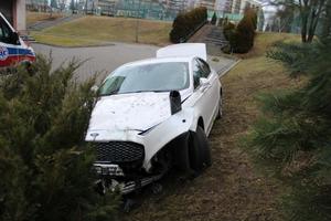 Biały samochód z uszkodzoną pokrywą silnika, stojący częściowo na trawniku, częściowo na chodniku