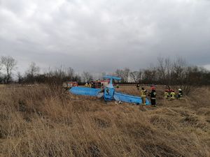 Widok od strony dziobu samolotu leżącego na grzbiecie. Obok grupa strażaków w tle samochody straży pożarnej.