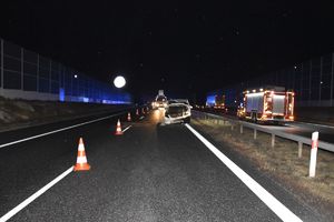 Zdjęcie białego mercedesa stojącego przy barierkach z uszkodzonym przodem. Po lewej stronie biało-czerwone pachołki wydzielające pas ruchu. zdjęcie wykonane nocą.