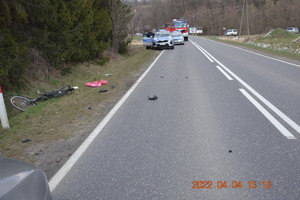 Widok miejsca wypadku rowerzysty. W tle radiowozy policyjne oraz straży pożarnej. W rowie melioracyjnym leży uszkodzony rower.