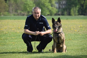 Umundurowany policjant kuca na trawie, a obok niego siedzi policyjny pies.