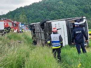 Przewrócony autokar wycieczkowy na polu zielony polu. Przed autokarem stoi policjant oraz strażak, kierujący działaniami ratowniczymi