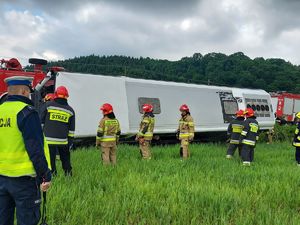 Przewrócony autokar, widok od strony jego dachu. Przed pojazdem stoją strażacy oraz policjanci.