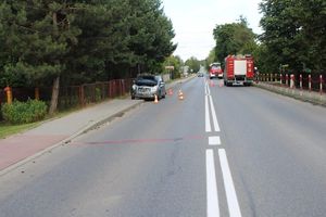 Mała toyota stojąca częściowo na chodniku. Widok z przodu pojazdu. otoczona pachołkami drogowymi. W tle pojazd straży pożarnej oraz strażak regulujący ruchem.