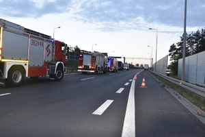 Miejsce wypadku. Po lewej stronie drogi, przy barierkach pojazdy straży pożarnej. Po prawej stronie wyznaczona strefa pachołkami drogowymi na końcu której stoi naczepa ciężarówki.
