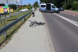 Na chodniku leży rower. W tle stojący w poprzek oznakowany radiowóz. Droga zablokowana.