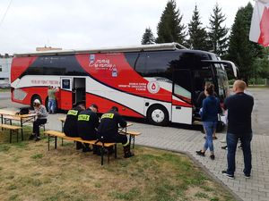 Na zdjęciu widoczny autobus z napisem MOBILNY PUNKT POBORU KRWI, obok stoją osoby w kolejce do krwiobusu, na ławce siedzi trzech strażaków