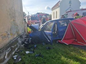 Uszkodzony przód niebieskiego samochodu osobowego, z tyłu za samochodem rozłożony czerwony parawan, na zdjęciu widoczni strażacy