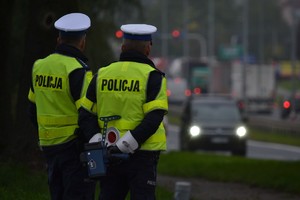 Dwóch policjantów ruchu drogowego stoi przy drodze, nadzoruje ruch. Jeden z policjantów trzyma za sobą ręczny miernik prędkości. W tle widać nadjeżdżające auto