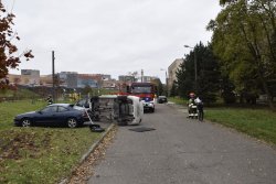 Widok ogólny miejsca kolizji. Przewrócony na prawym boku biały samochód oraz niebieski pojazd stojący na parkingi, na jego bagażniku przewrócona rurka metalowa.