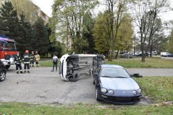 Widok z przodu samochodu Toyota. Z tyłu przewrócony biały Peugeot leżący na prawym boku. Z lewej strony pojazdy straży pożarnej obok stojący strażacy