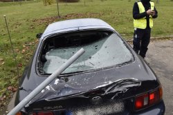 Zbliżenie na uszkodzenia Toyoty. Wybita tylna szyba oraz uszkodzona pokrywa bagażnika. Widoczna rurka znaku drogowego. w tle stojący policjant.