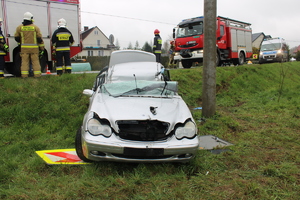 Zdjęcie przedstawiające przód mercedesa klasy C stojącego na kołach na trawie obok betonowego słupa energetycznego. Widoczne uszkodzenia dachu pojazdu i zniszczona przednia szyba