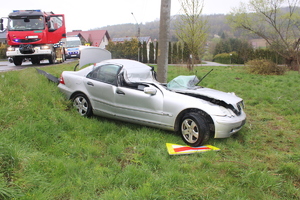 Zdjęcie przedstawiające prawy bok mercedesa klasy C stojącego na kołach na trawie przed betonowym słupem energetycznym. Widoczne uszkodzenia dachu pojazdu i zniszczona przednia szyba.