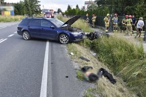 Miejsce wypadku drogowego. Na pierwszym planie rozrzucone elementy oraz kask motocyklisty, dalej niebieski samochód osobowy stojący w poprzek drogi